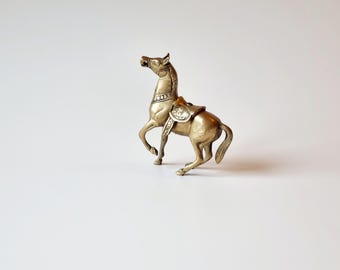May include: A gold-colored metal horse figurine with a saddle and intricate details. The horse is standing on a white surface.
