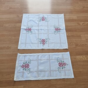 以下が含まれることがあります： グリッドパターンと赤と緑の花柄が施された白い刺繍のテーブルクロス2枚。大きなテーブルクロスは正方形で、小さいテーブルクロスは長方形です。