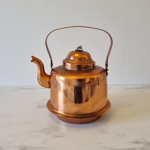 Könnte beinhalten: Ein kupferfarbener Wasserkocher mit gebogenem Ausguss, gewölbtem Deckel und Griff. Der Wasserkocher steht auf einem runden Sockel. Der Griff wölbt sich über die Oberseite. Der Wasserkocher ist aus glänzendem Metall.
