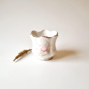 May include: A small white ceramic planter with a scalloped rim and a floral design featuring pink roses. The planter has a gold trim around the rim.