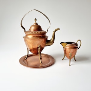 Puede incluir: Una tetera de cobre con asa de latón y tres patas, sentada sobre una bandeja de cobre. Una jarra de crema de cobre a juego con asa de latón y tres patas está al lado de la tetera.