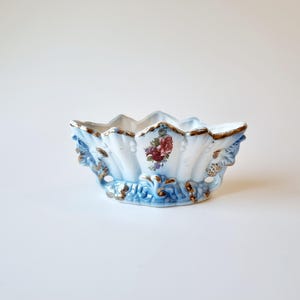 Può includere: Una ciotola in ceramica bianca con accenti blu e dorati. La ciotola ha un bordo a forma di conchiglia e un motivo floreale.