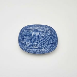 May include: A blue and white ceramic plate with a detailed illustration of a town scene. The plate has a scalloped edge and a raised rim. The date "1993" is printed on the bottom of the plate.