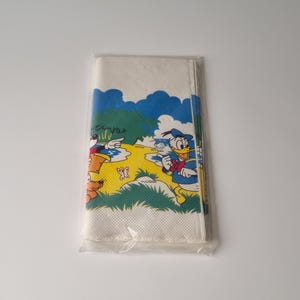 Pode incluir: Um pacote de guardanapos brancos com uma cena de desenho animado colorida com o Pato Donald e outros personagens. O design inclui uma paisagem amarela e verde com céu azul e nuvens. Os guardanapos são selados em uma embalagem plástica transparente.
