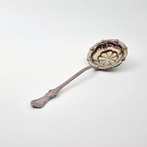 Puede incluir: Un colador de té de plata con un mango decorativo y un cuenco perforado en forma de flor.