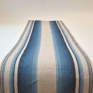 Könnte beinhalten: Ein rechteckiger, gewebter Tischläufer mit vertikalen Streifen in verschiedenen Blau- und Off-White-Tönen. Das Design des Läufers weist einen zentralen hellblauen Streifen auf, der von dunkleren blauen und off-White-Streifen flankiert wird, wodurch ein Gradienteneffekt entsteht.