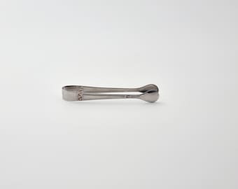 Puede incluir: Un par de pinzas para azúcar de metal plateado con acabado pulido. Las pinzas cuentan con un mango en forma de U conectado a dos extremos redondeados en forma de cuchara. Hay un pequeño patrón geométrico decorativo grabado cerca de la bisagra.