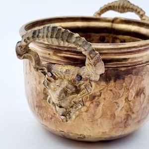 Peut inclure: Un pot en laiton martelé avec deux poignées en tête de bélier. Le pot a une surface texturée et est de couleur or chaud.