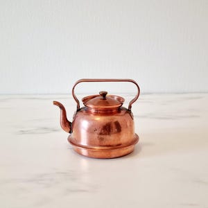 Puede incluir: Una tetera de cobre pequeña con cuerpo redondeado y tapa. La tetera tiene asa y pico.