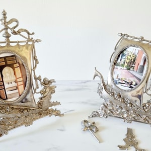 May include: Two silver-toned picture frames shaped like ships with ornate details. Each frame has an oval opening and a photo inside. The frames are on a white surface.