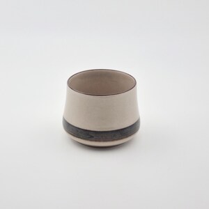 May include: A white ceramic cup with a brown and black band around the middle.