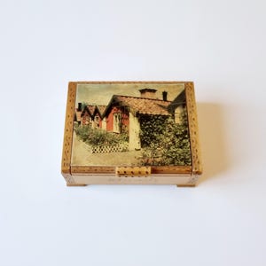 Può includere: Una scatola di legno con un coperchio incernierato. Il coperchio presenta una fotografia vintage di una fila di piccole case con tetti rossi e vegetazione verde.