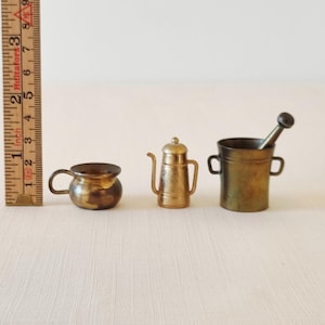 Puede incluir: Juego de miniaturas de utensilios de cocina de latón que incluye una olla pequeña, una cafetera y un mortero con mano. La olla tiene aproximadamente 2,5 cm de diámetro. La cafetera mide aproximadamente 4 cm de alto. El mortero y la mano miden aproximadamente 4,5 cm de alto.