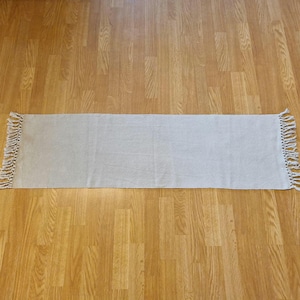 May include: A rectangular table runner in a solid light beige color. The fabric features a woven texture throughout the surface. Both short ends of the runner are finished with decorative, hand-knotted fringe tassels made of the same material.