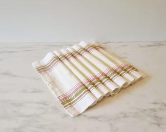 May include: A stack of folded white linen napkins with stripes in pink and green. The napkins are arranged on a marble surface, with the stripes running horizontally across the fabric. The napkins are likely for a kitchen or dining setting.