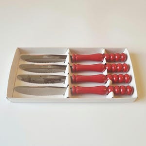 May include: A set of four stainless steel serrated knives with bright red, rounded, bead-textured handles. The knives are arranged horizontally in a white cardboard tray, featuring metallic silver blades and vibrant red, cylindrical, segmented grips.