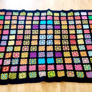 May include: A colourful crocheted blanket with a black background. The blanket is made up of many small squares, each with a different colour. The colours are bright and vibrant, and the blanket is a great way to add a pop of colour to any room.