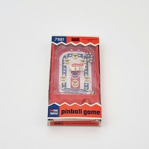 Puede incluir: Una caja de cartón roja y azul con un mini juego de pinball dentro. La caja tiene las palabras "pinball game" y "MMG" impresas. El juego tiene un diseño rojo y blanco con una etiqueta de precio que dice "KONBERG 6.85".