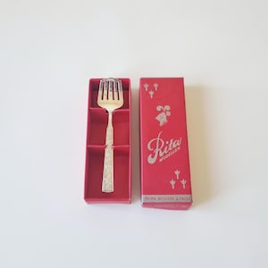 May include: A silver serving fork with a decorative handle rests in a red box. The box is open, revealing the fork's tines and handle. The box has the text "Rita Modeller" in silver script and "Palma Nysilver Alpaca" printed on the side.