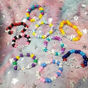 Könnte beinhalten: Eine Sammlung bunter Perlenarmbänder mit verschiedenen Texten. Die Armbänder weisen eine Vielzahl von Farben auf, darunter Rot, Schwarz, Weiß, Rosa, Blau, Gelb und Lila. Einige Armbänder buchstabieren Wörter wie "BEE", "HUSK", "ANGEL", "ROX", "XIAO", "SMILE" und "VOXTECH".