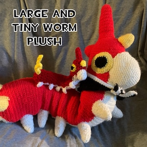 May include: A handmade crochet worm plush in red, white, black, and yellow. The plush has a large head and body with a smaller worm attached. The text "LARGE AND TINY WORM PLUSH" is displayed in the image.