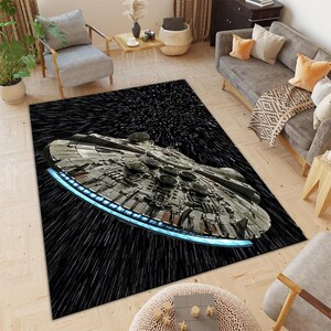May include: A rectangular area rug featuring a detailed illustration of the Millennium Falcon spacecraft against a black background with white streaks, creating a sense of speed. The rug has a blue glowing edge.