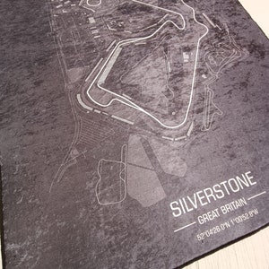 May include: A black and white map of the Silverstone racetrack in Great Britain. The map shows the layout of the track and surrounding area. The text "SILVERSTONE - GREAT BRITAIN - 52&deg;04'26.0"N 1&deg;00'52.8"W" is printed below the map.