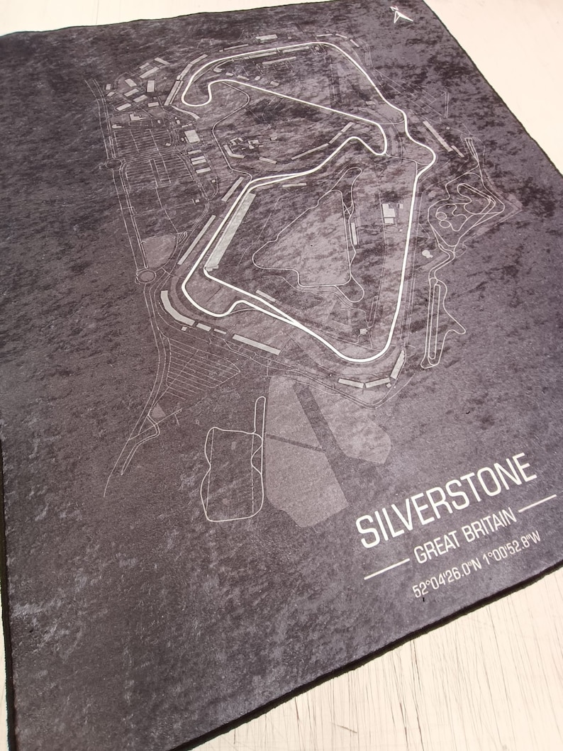 May include: A black and white map of the Silverstone racetrack in Great Britain. The map shows the layout of the track and surrounding area. The text "SILVERSTONE - GREAT BRITAIN - 52&deg;04'26.0"N 1&deg;00'52.8"W" is printed below the map.