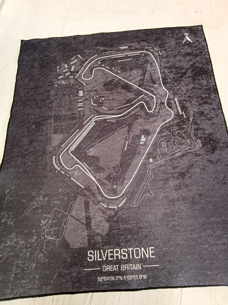 May include: A black and white map of the Silverstone racetrack in Great Britain. The map shows the layout of the track and surrounding area. The text "SILVERSTONE" and "GREAT BRITAIN" is printed in white at the bottom of the map. The coordinates "52&deg;04'26.0"N 1&deg;00'52.8"W" are printed below the text.