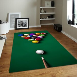 May include: A rectangular green rug with a printed image of a pool table. The rug features a rack of billiard balls in various colors, a white cue ball, and a pool cue. The rug is placed on a wooden floor.
