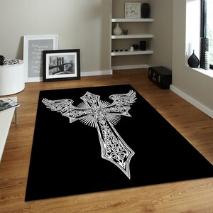 May include: A rectangular area rug featuring a solid black background with a large, centred white graphic design. The design depicts an ornate cross with intricate scrollwork patterns and stylised wings extending from the upper horizontal arms.