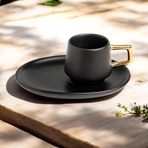 Op de afbeelding: Een zwarte keramische koffiekop met een gouden oor staat op een bijpassende zwarte schotel. De kop en schotel staan op een houten oppervlak, met natuurlijk licht en schaduwen.