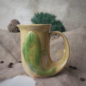 Może przedstawiać: Ręcznie robiony ceramiczny kubek o unikalnym wzorze. Kubek jest w jasnym beżowym kolorze z dużym zielonym abstrakcyjnym kształtem z przodu. Ma zakrzywiony uchwyt i stoi na jutowej powierzchni. Widoczne są również ziarna kawy i szyszka.