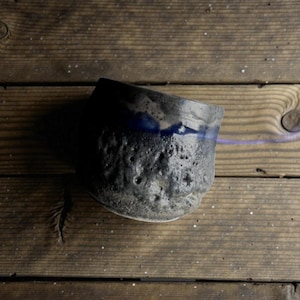 May include: A handmade ceramic bowl with a textured, dark gray exterior and a blue interior rim. The bowl sits on a wooden surface, showcasing its unique, organic shape and design. The bowl's surface has a rough, pitted texture.