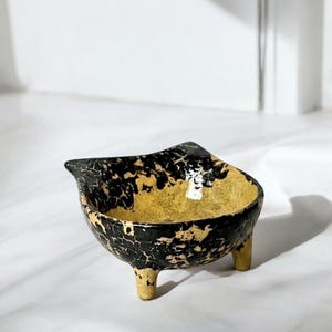 Könnte beinhalten: Handgefertigte Keramikschale mit einzigartigem Design. Die Schale hat eine schwarze, craquelierte Außenseite über einem goldenen Innenraum. Sie steht auf drei kleinen Beinen und hat eine asymmetrische Form, ca. 15 cm Durchmesser.