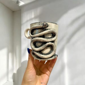 Puede incluir: Taza de cerámica color crema con borde y asa negros. Un diseño de serpiente texturizada y enrollada envuelve el exterior de la taza. La cabeza de la serpiente está en la parte superior, cerca del borde. La taza es sostenida por una mano con esmalte de uñas azul.