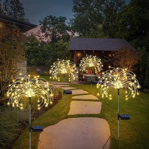 May include: Four solar powered firework lights illuminate a pathway in a garden. The lights are made of thin metal wires with small white LED lights that create a firework effect. The lights are on stakes that are inserted into the ground.