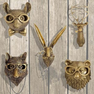 Può includere: Cinque sculture dorate di teste di animali con occhiali e peli facciali. Le sculture sono montate su uno sfondo di pannello di legno. Gli animali includono un coniglio, un cervo, un orso e altri due animali.