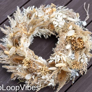 Pode incluir: Uma grinalda circular composta por flores e gramíneas secas em tons de bege, creme e castanho. A grinalda apresenta diversas texturas e formas, criando uma estética natural e rústica. Um elemento decorativo para a decoração da casa.