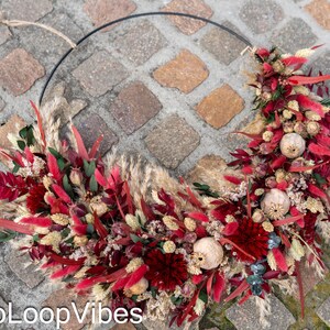 Może przedstawiać: Okrągły wieniec z suszonych kwiatów i traw w odcieniach czerwieni, bordo i beżu. Wieniec ułożony jest na czarnym metalowym pierścieniu i zawiera torebki nasienne. Tekst "DecoLoopVibes" jest widoczny w lewym dolnym rogu.