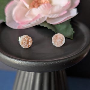 Pode incluir: Dois brincos redondos com uma superfície texturizada de cor pêssego claro. Os brincos são exibidos em um pedestal decorativo cinza escuro, com uma grande flor rosa e branca ao fundo. Joias.