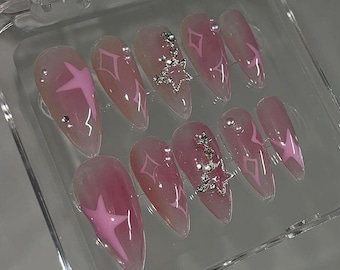 Puede incluir: Un conjunto de diez uñas postizas rosas con un diseño de estrella blanca y pedrería plateada.