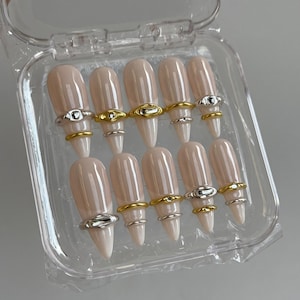 May include: A set of ten almond-shaped press-on nails in a glossy nude beige finish, each adorned with a single metallic ring accent in either gold or silver tones, presented in a clear plastic storage container with a hinged lid.