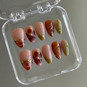 May include: A set of ten false nails with a fall-themed design. The nails are nude with gold accents and brown and green leaf designs.