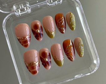 Puede incluir: Un conjunto de diez uñas postizas con un diseño de otoño. Las uñas son de color nude con detalles dorados y diseños de hojas marrones y verdes.