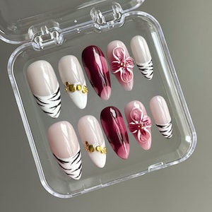 May include: A set of ten false nails in a clear plastic case. The nails are almond shaped and have a variety of designs. Some nails are painted a deep burgundy color with a white tip and a zebra print design. Other nails have a white base with gold accents and a 3D pink flower design.