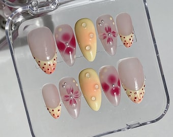 May include: A set of ten false nails in a clear plastic case. The nails are almond shaped and have a light pink base. Some nails have a red and white polka dot design, while others have a pink flower design with a pearl accent.