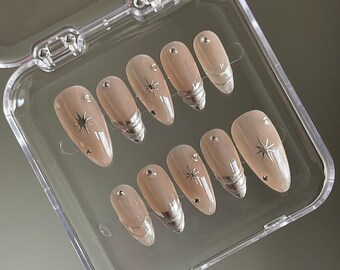 May include: A set of ten nude-colored false nails with silver tips and a star design. The nails are arranged in a clear plastic case.