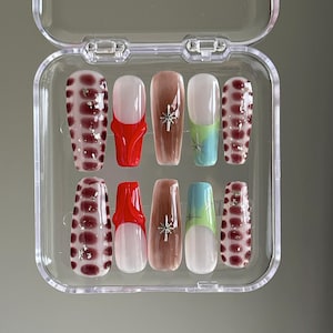 May include: A set of ten false nails in a clear plastic case. The nails are in various designs, including red, brown, white, and blue. Some nails have a snowflake design, while others have a polka dot design.