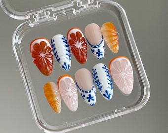 May include: A clear plastic box with a set of ten fake fingernails. The nails are decorated with a white and blue floral design and orange accents.